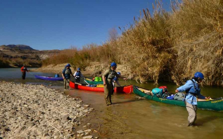 a group of gap year students walk beside canoes, guiding them through a shallow portion of water on an outward bound expedition 
